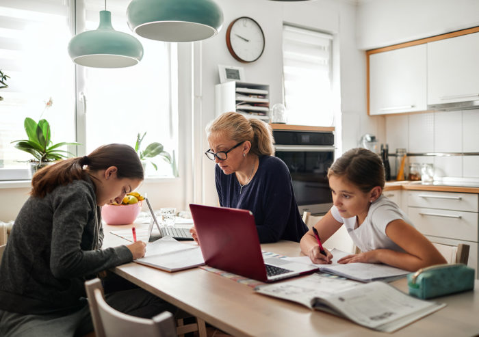 Woman working from home at table with two daughters in grade school in remote learning and doing homework.