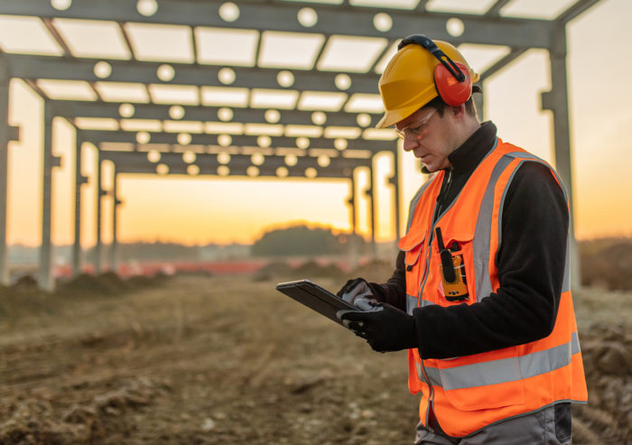 Constructor worker or contractor in field in front of steel framed building using tablet.