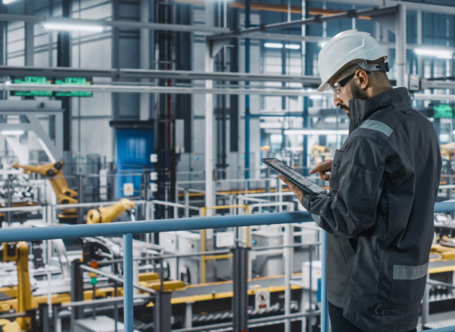 Engineer in hardhat overseeing robotic arms in a production line while reviewing analytics on customized connected tablet